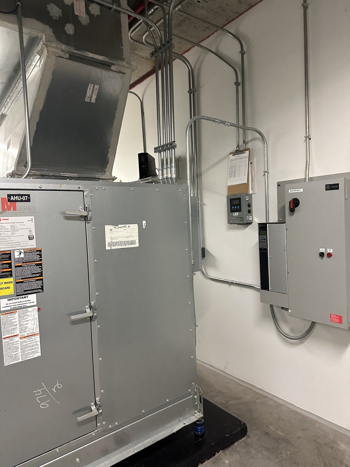 A Trane M-Series air handler unit in a commercial mechanical room with a wall-mounted VFD and disconnect