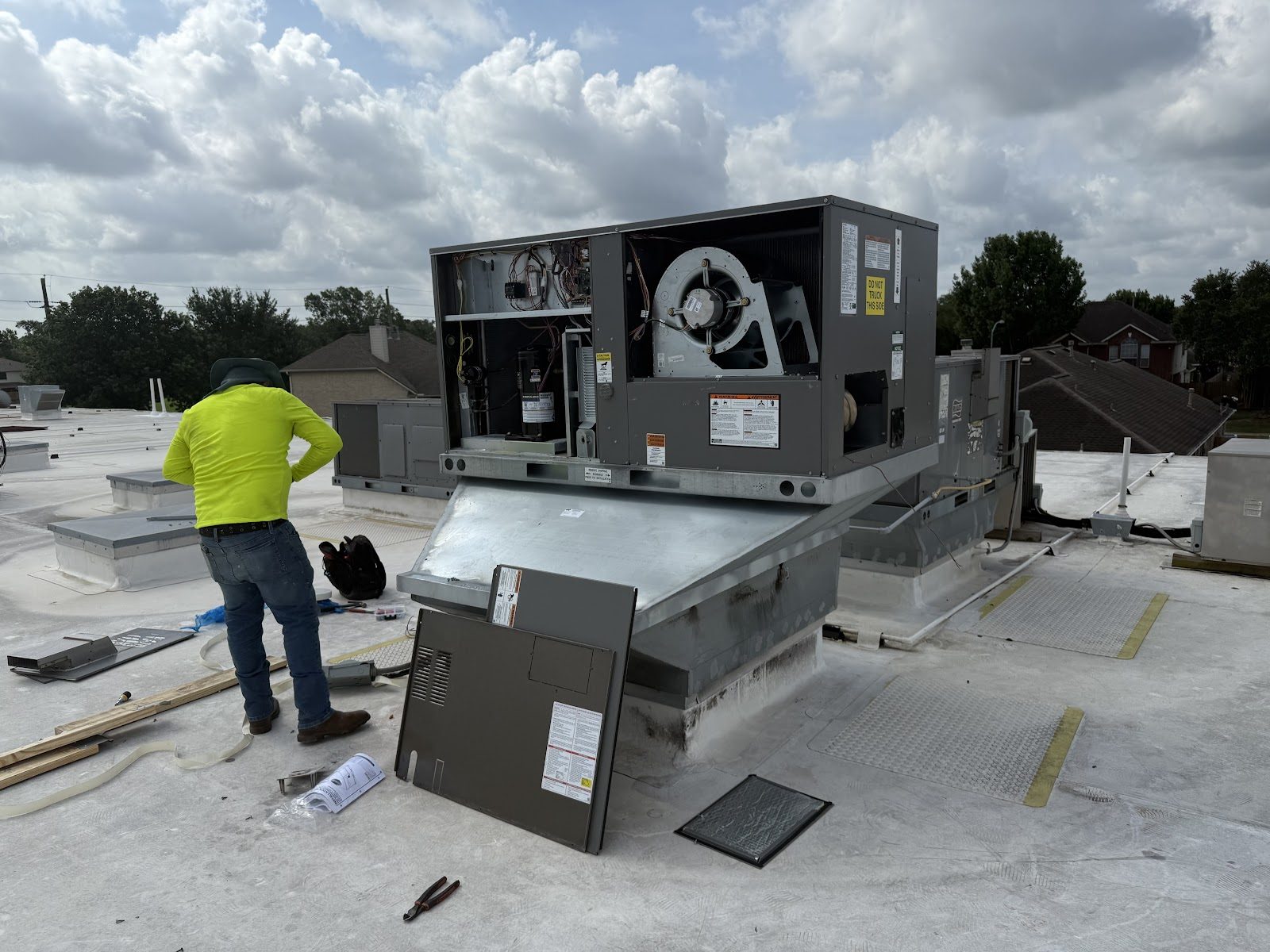 A Prime HVAC Solution technician in a hi-vis shirt working inside an opened rooftop packaged unit under a cloudy Texas sky